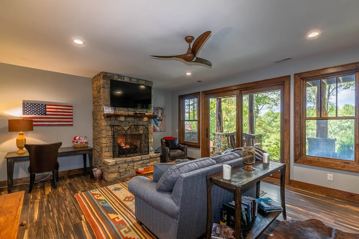 A warm, inviting lower-level living area featuring a cozy gas fireplace, a comfortable sofa, large windows with scenic views, a decorative rug, and an American flag on the wall. The space is arranged for relaxing, socializing, and enjoying the ambiance of the fire.