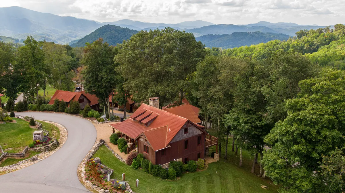 The image shows a beautiful single-family house with a striking red roof nestled among lush trees, with a sweeping mountain backdrop and a curved road leading to the property. The setting is tranquil, with natural landscaping and panoramic views that showcase the serene beauty of the North Carolina mountains.