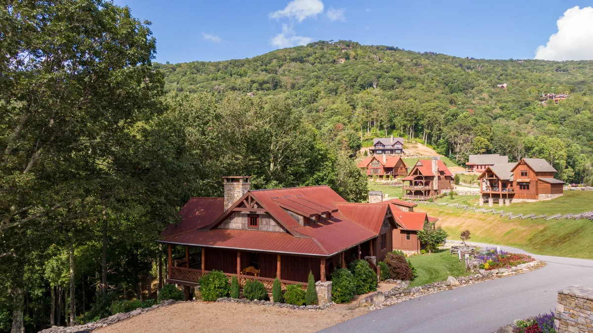 A rustic cabin with a red metal roof surrounded by trees, distant hills, and neighboring cabins. The setting is cozy yet expansive, ideal for outdoor family barbecues, nature walks, and group activities.