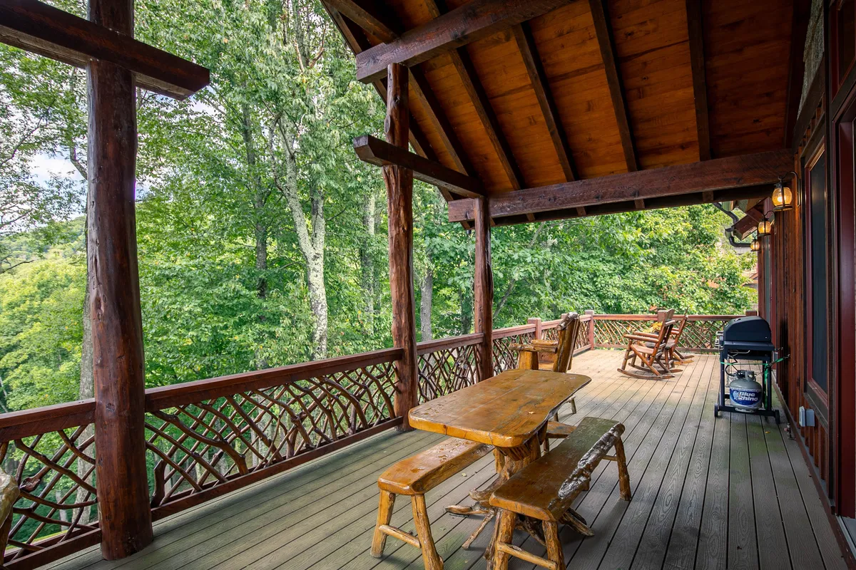 An outdoor deck scene with a wooden table set for dining, classic rocking chairs, and beautiful natural surroundings. The positioning suggests the perfect spot for sharing meals or enjoying a drink while watching the sunset.