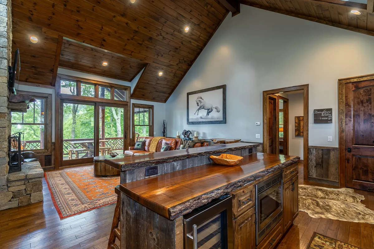 This image offers a sweeping view of the open-concept living area, seamlessly transitioning into the kitchen. The stone fireplace, horse artwork, large windows with scenic views, and a wooden kitchen island are all visible. The arrangement encourages social interaction, with ample seating and spaces for both relaxation and culinary activity.
