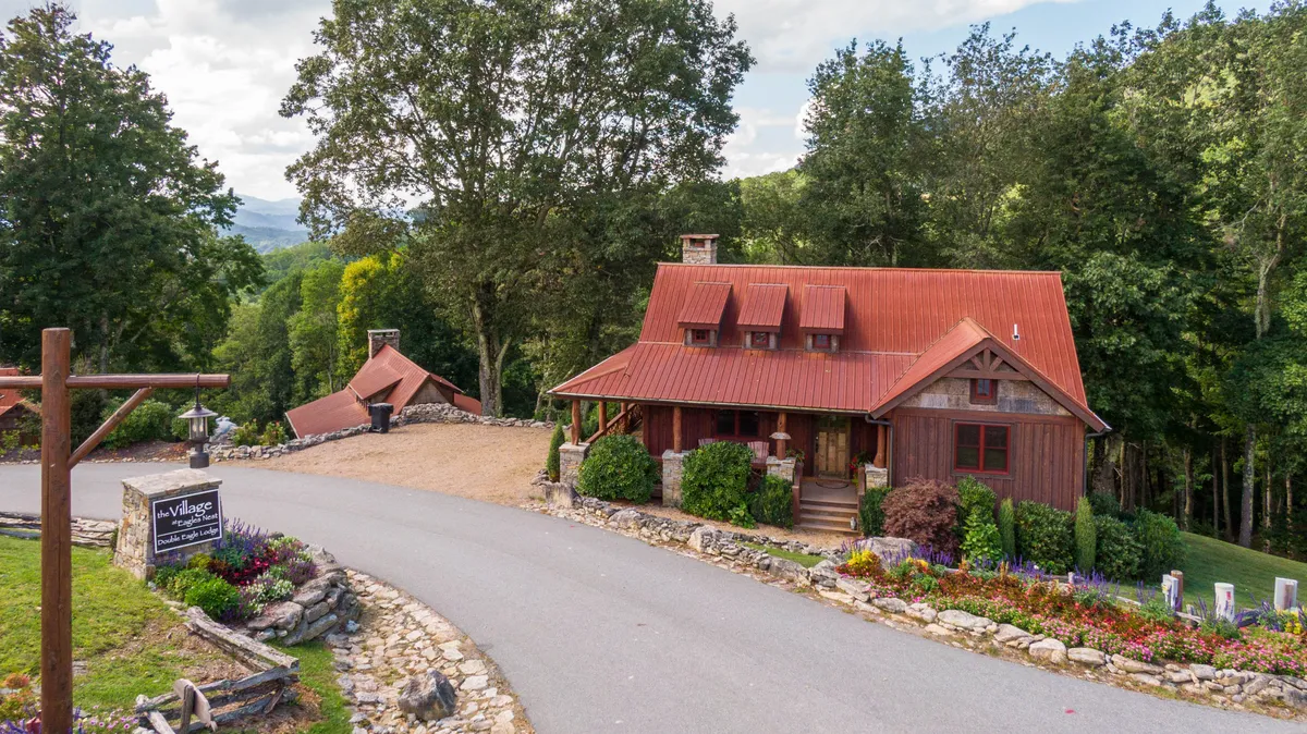 A welcoming view of a rustic house with a red roof, surrounded by colorful stone pathways, flower beds, and a signpost. The porch and garden area suggest leisurely strolls, outdoor gatherings, and access to trails and outdoor amenities within the property’s grounds.