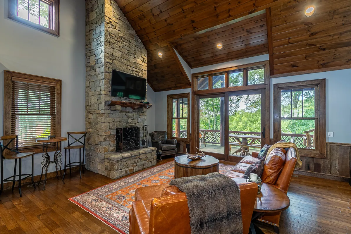 A spacious living room with a stunning stacked stone wood-burning fireplace forms the focal point. Large windows let in natural light and offer glimpses of the outdoors, while leather seating and a decorative rug create a cozy atmosphere. The soaring wooden ceiling accentuates the sense of space and comfort, perfect for gatherings around the fire or enjoying a game night.