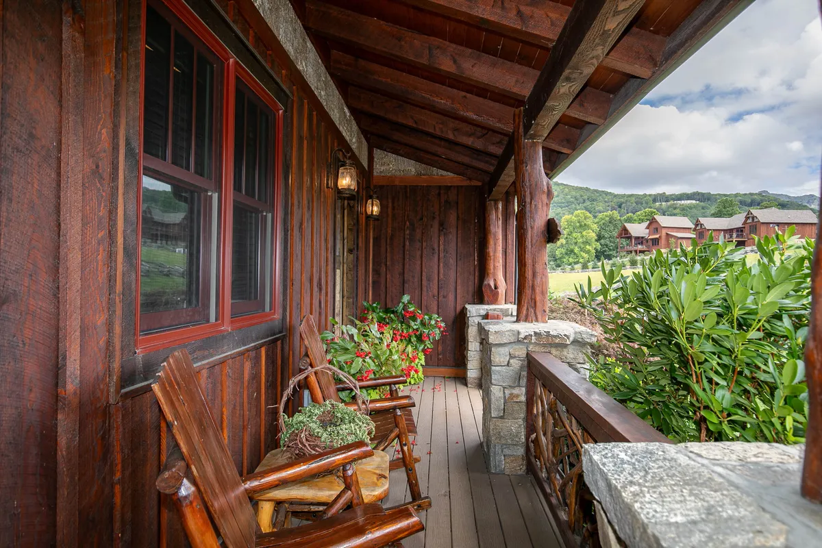 A cozy outdoor porch scene with wooden rocking chairs, inviting stone railing, and vibrant flowering plants. The large windows in the background suggest seamless indoor-outdoor living, while the scene evokes peaceful moments spent enjoying sunset views or quiet conversation.