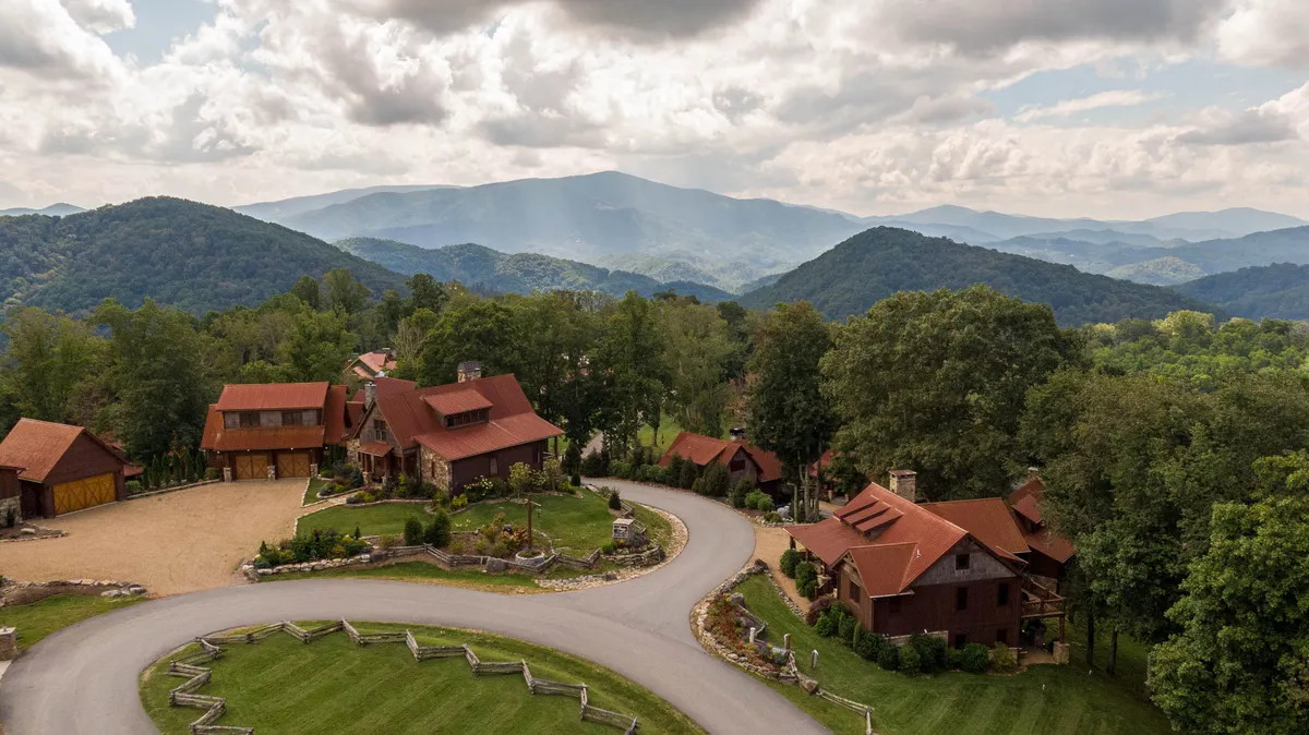 A panoramic view of a mountain range with a circular driveway, wooden cabins nestled amidst lush greenery, and wide open spaces ideal for family gatherings and exploration. The scene conveys tranquility and the scenic beauty of the resort’s natural surroundings.