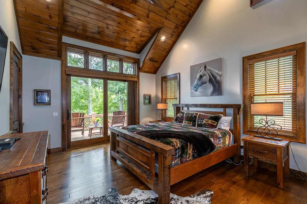 This image features a wooden bed adorned with plush linens, set against large windows that flood the room with natural light. Side tables with lamps and rustic horse artwork add to the tranquil and rustic vibe of the bedroom, with subtle hints of deck access in the background, inviting relaxation and connection to nature.