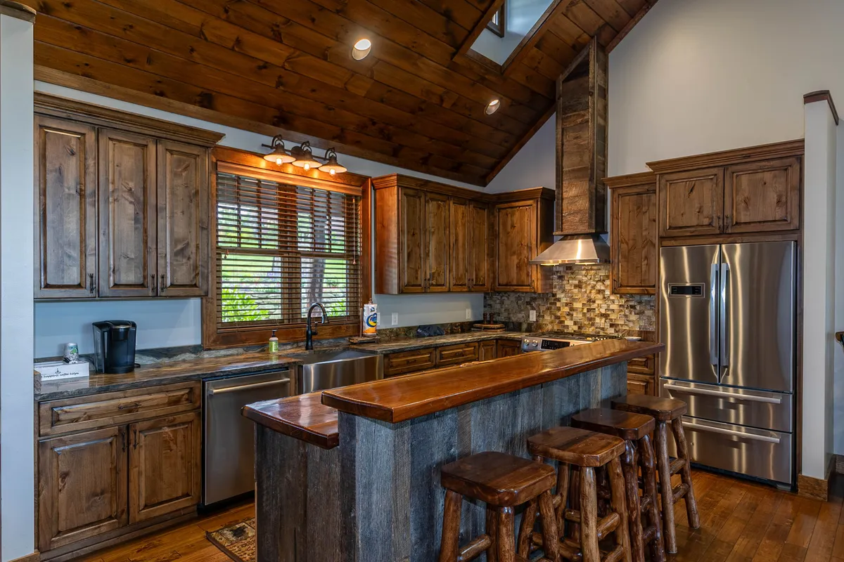 A close-up focus on the gourmet kitchen area featuring a large wooden island with bar stools, stone backsplash, and modern appliances. The layout encourages casual dining, coffee conversations, and participation in meal preparation. Warm wood tones and cozy lighting add to the communal ambiance.