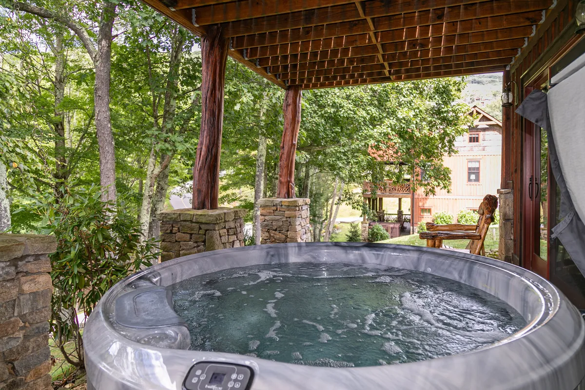 An exterior scene featuring a private hot tub nestled against stone walls and wooden beams, surrounded by lush natural scenery. The space exudes tranquility and is perfect for enjoying serene relaxation while soaking in the hot tub, with views of the trees and landscape.